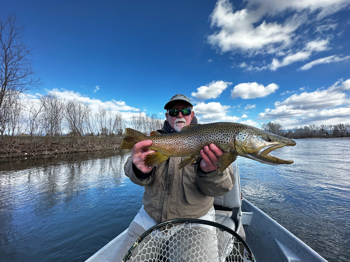 Fisherman holding large brown trout seated in boat on river under partly cloudy blue sky with leafless trees nearby