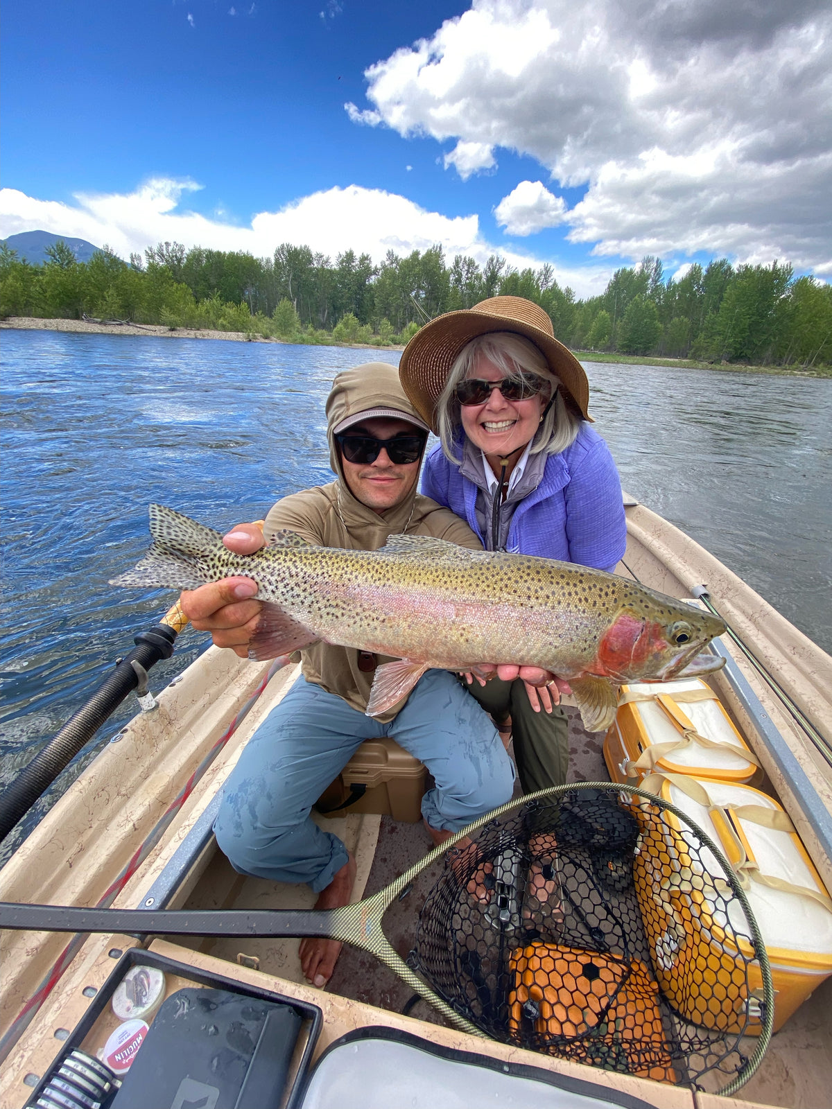 Two anglers smiling in a boat holding a large rainbow trout with trees and cloudy sky in the background