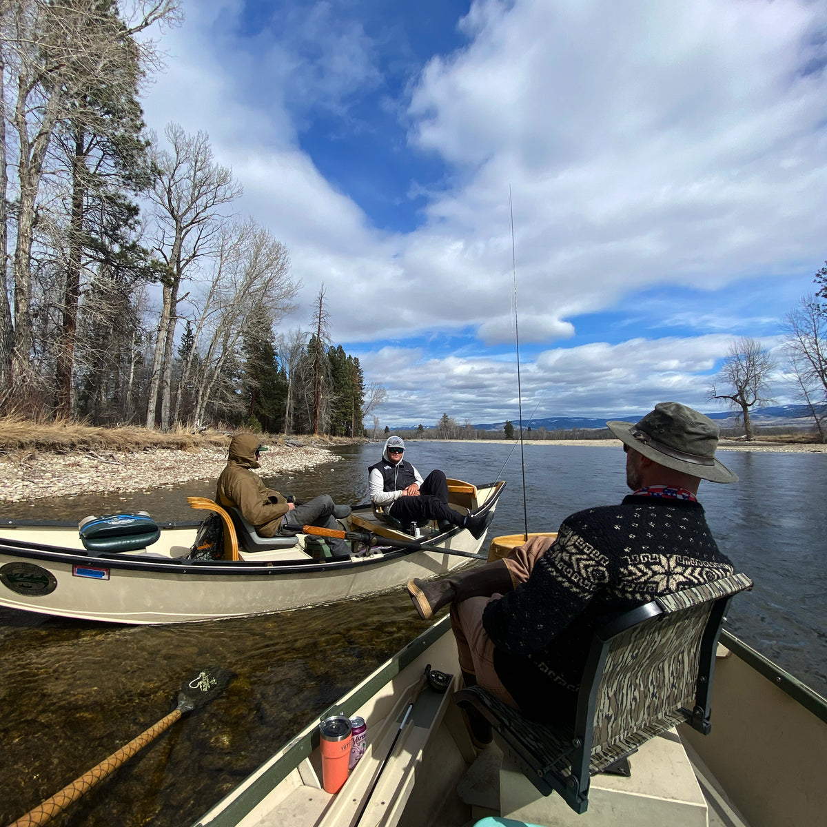 Three anglers sitting in separate boats on a calm river under a partly cloudy sky near leafless trees