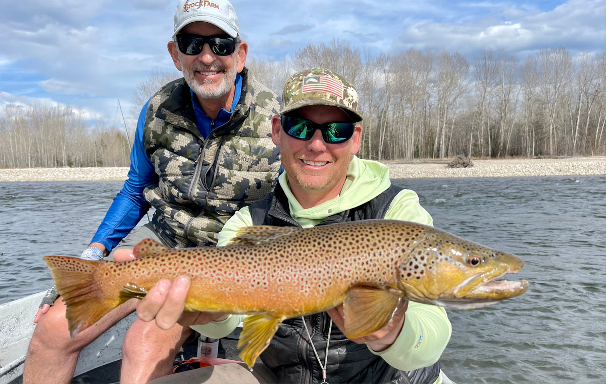 Two men in sunglasses holding a large brown trout on a river with leafless trees and partly cloudy sky behind